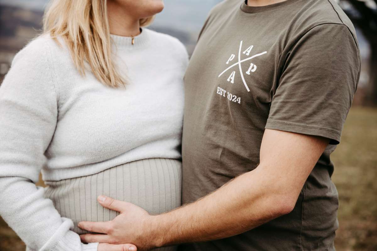Schwangere und ihr Partner im PAPA-Shirt halten gemeinsam den Babybauch