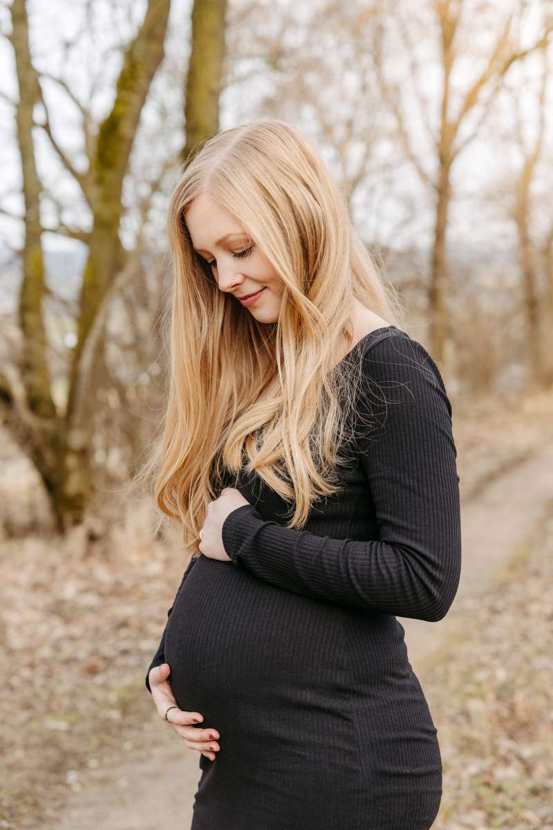 Schwangere im schwarzen Kleid h&auml;lt ihren Bauch, dahinter kahler Wald