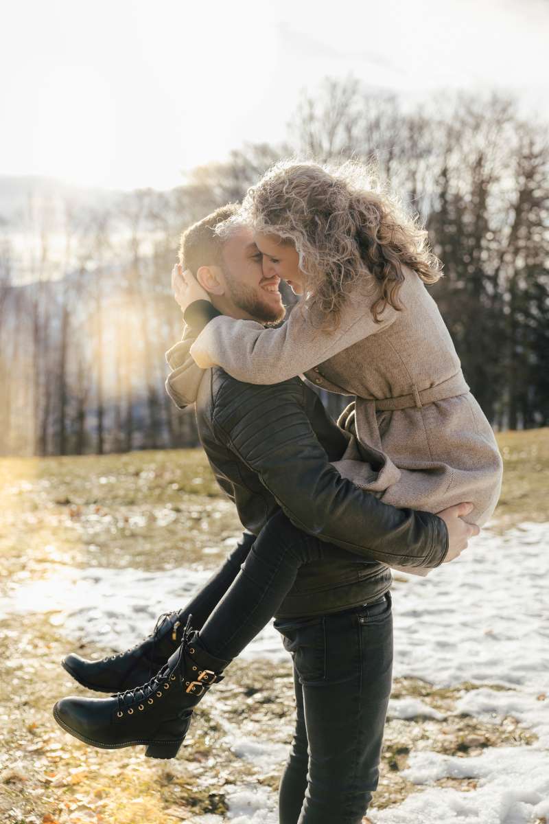 Mann hebt seine Partnerin lachend hoch, im Hintergrund Wald und Restschnee